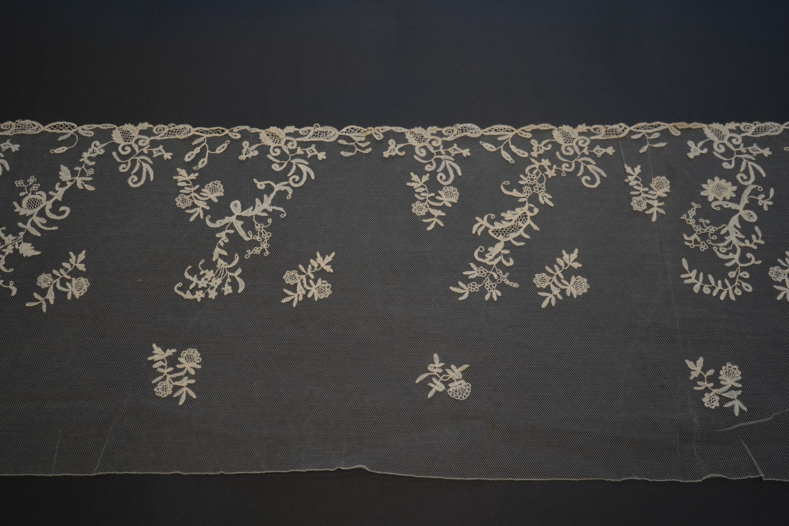 A 19th century five yard flounce of Brussels bobbin appliqué lace applied onto fine machine net, worked in a trailing floral design with an ornate border and sprig motifs, 457.5 cm long x 36cm deep. Condition - possibly